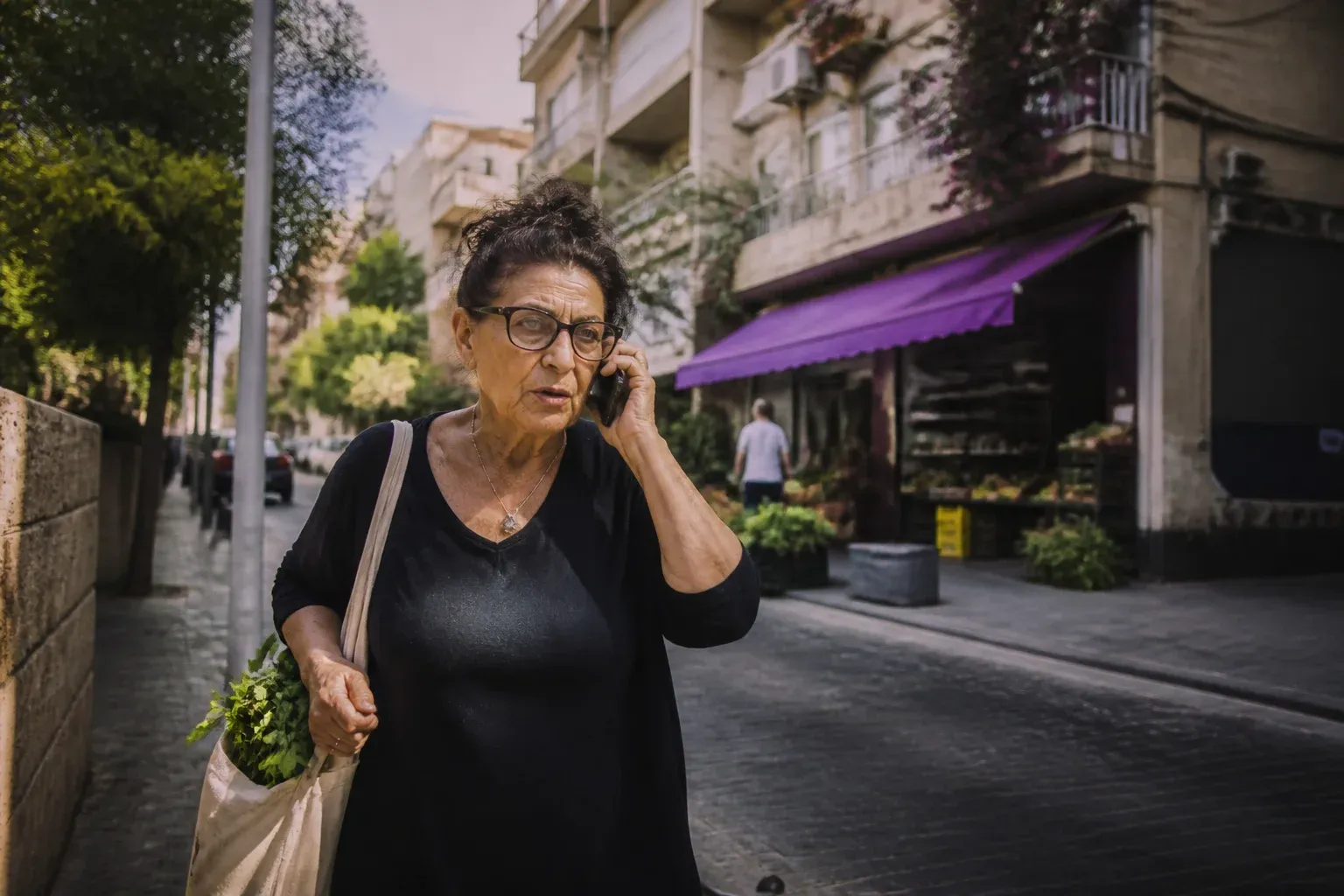 אישה בת שישים מדברת בטלפון ברחוב שכונתי עם שקית קניות