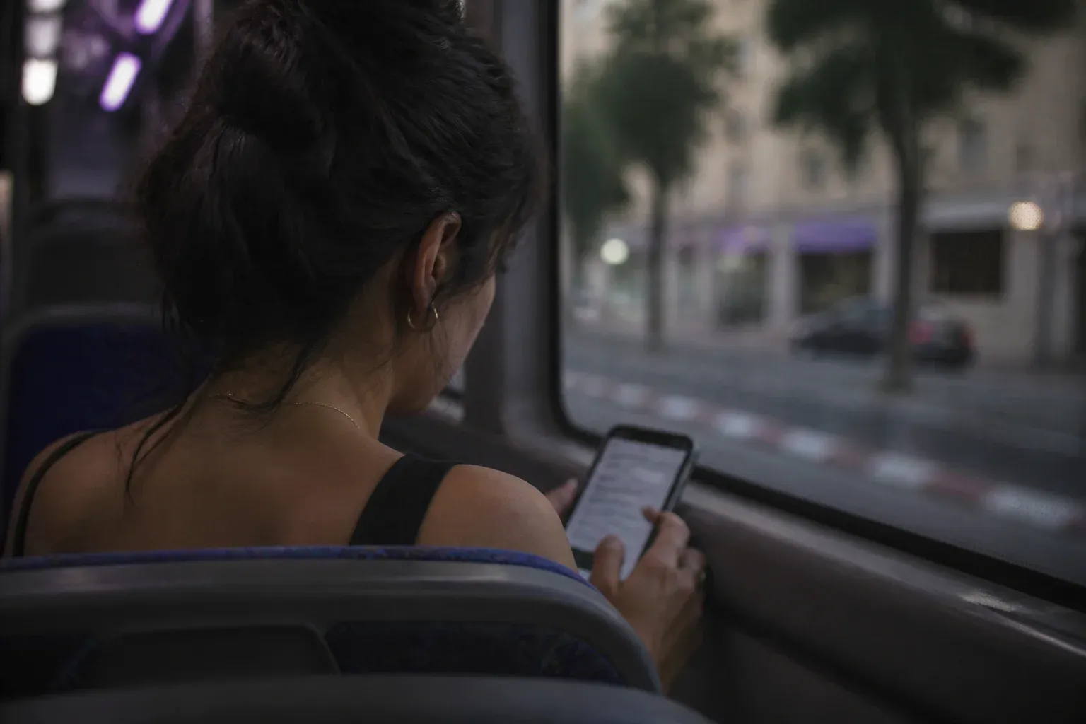 אישה באוטובוס מקישה בטלפון על טופס מטושטש כשהעיר חולפת בחלון
