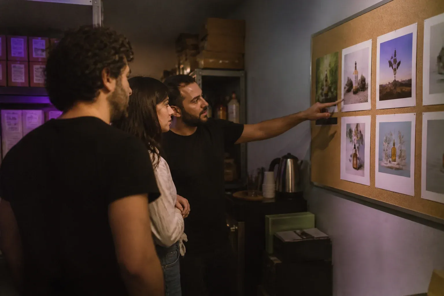 שלושה אנשים בחדר אחורי של חנות בודקים הדמיות מודעה מודפסות על לוח שעם